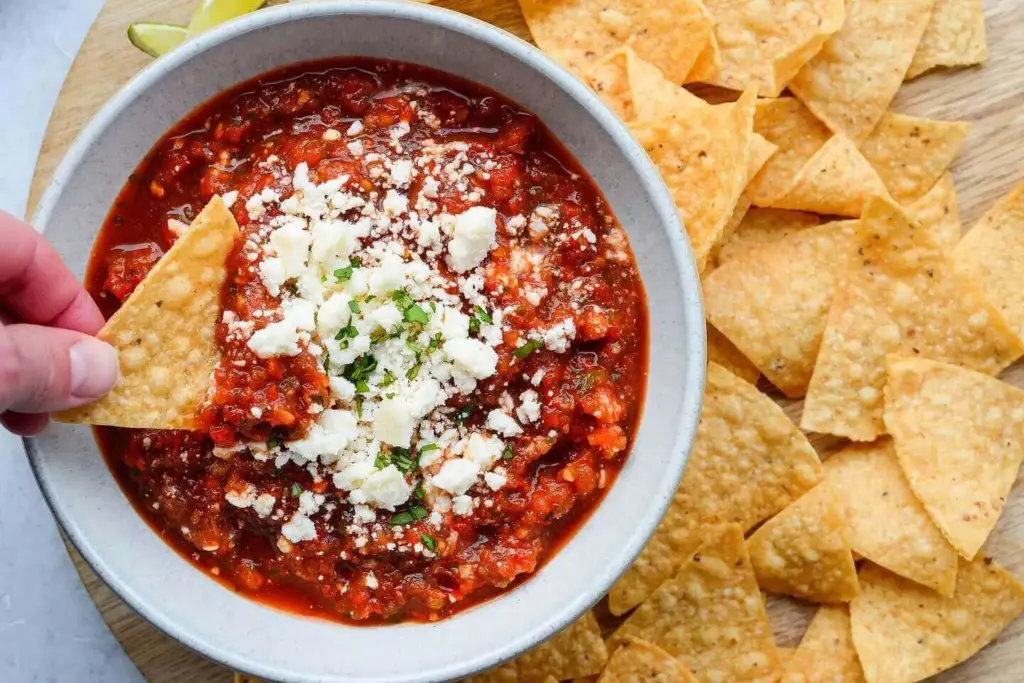 A hand dips a tortilla chip into a bowl of red salsa topped with crumbled white cheese. More chips are scattered on the side.