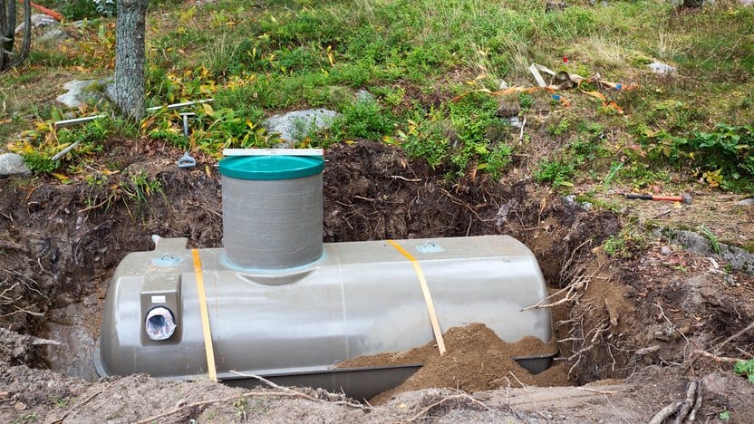 A partially buried plastic septic tank with a green lid is installed in a dug-out earth pit, surrounded by soil and plants.