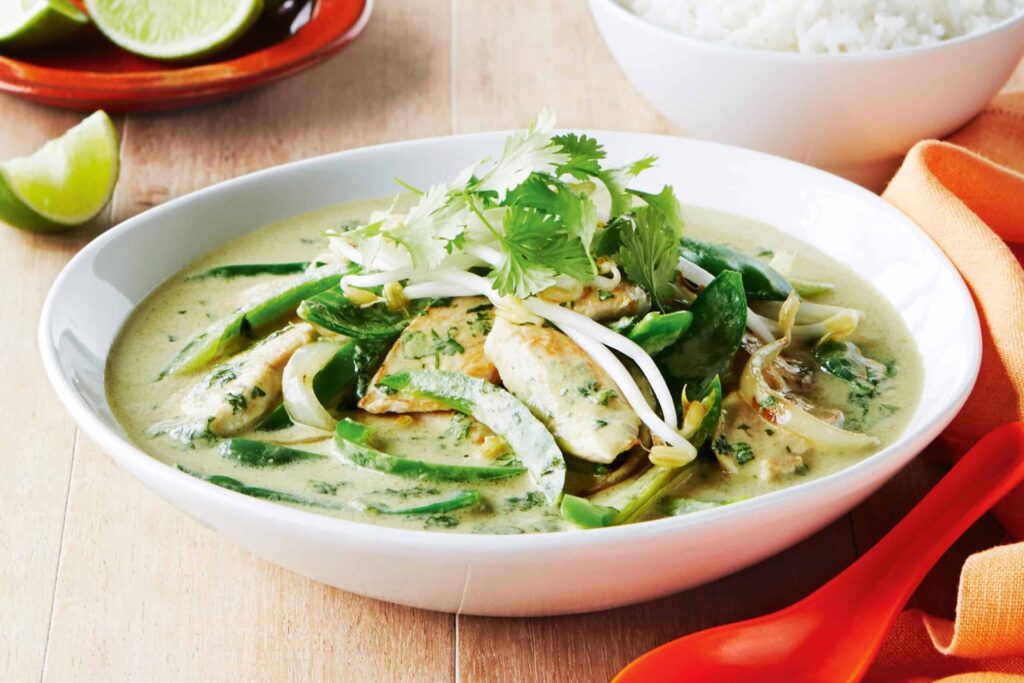 A bowl of green curry with chicken, snow peas, bean sprouts, and cilantro, accompanied by a bowl of white rice and lime wedges on a wooden table.