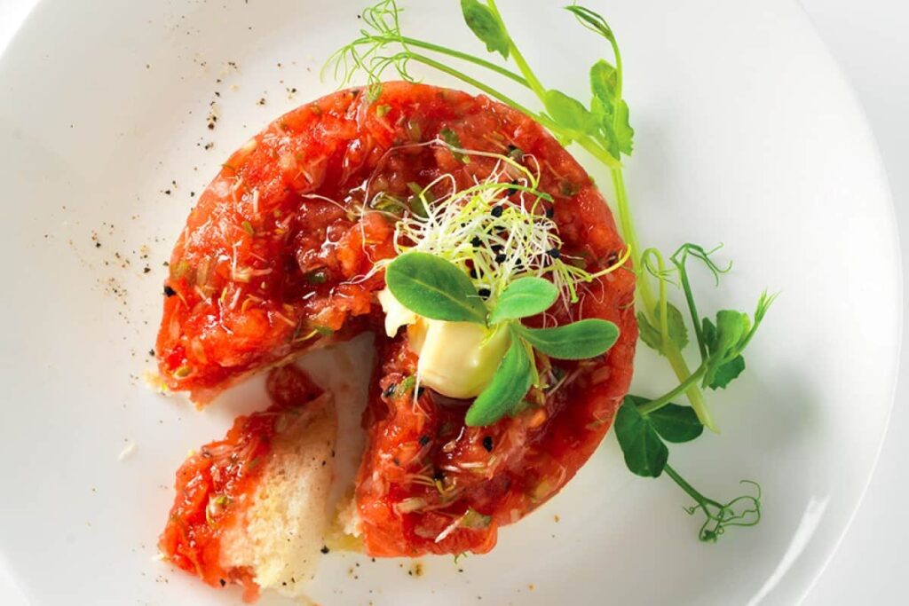 A round serving of tomato tartare garnished with herbs and sprouts on a white plate, with a small piece missing and a slice of bread on the side.