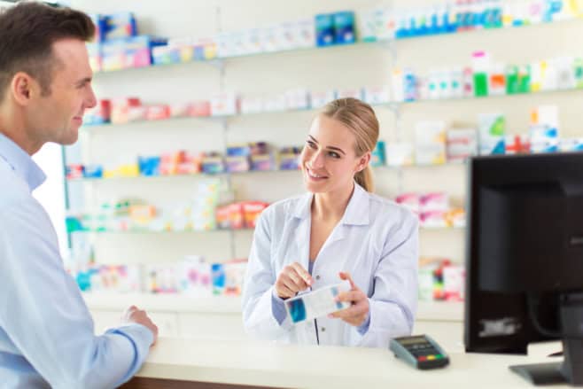 A pharmacist in a white coat assists a customer at a pharmacy counter, surrounded by shelves of medicine.