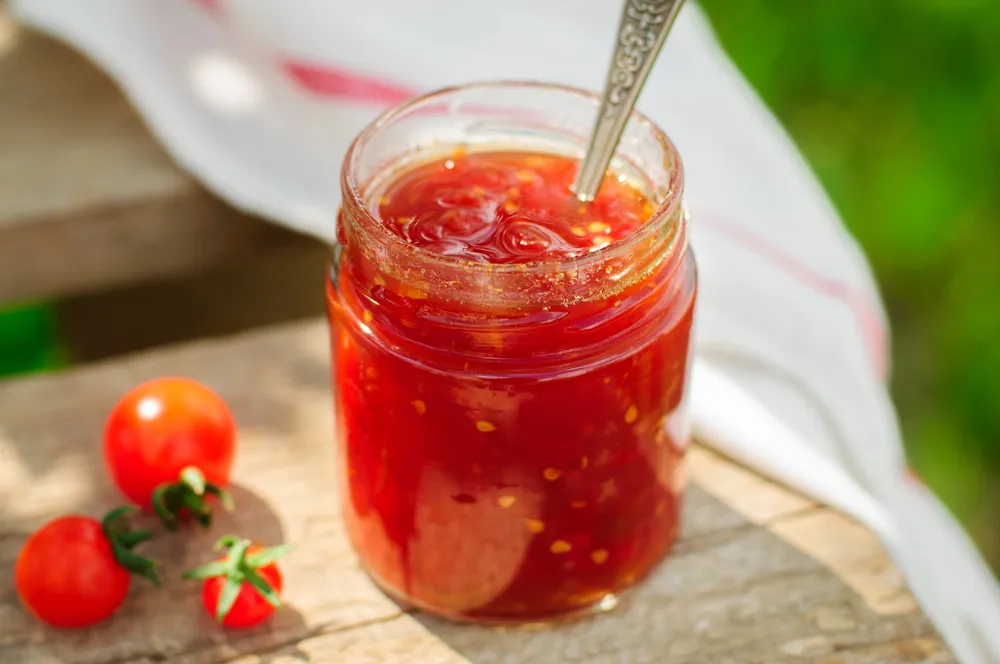 A jar of tomato jam with a spoon inside, placed on a wooden surface next to a few fresh tomatoes.