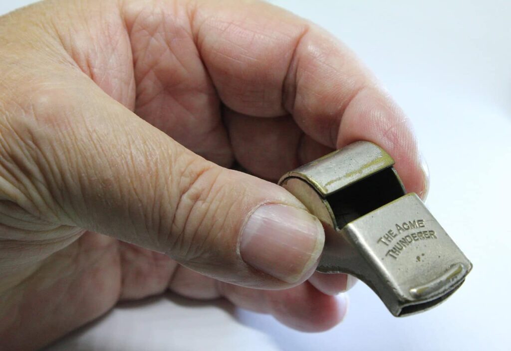 A hand holding a metal whistle with "The Acme Thunderer" engraved on it.