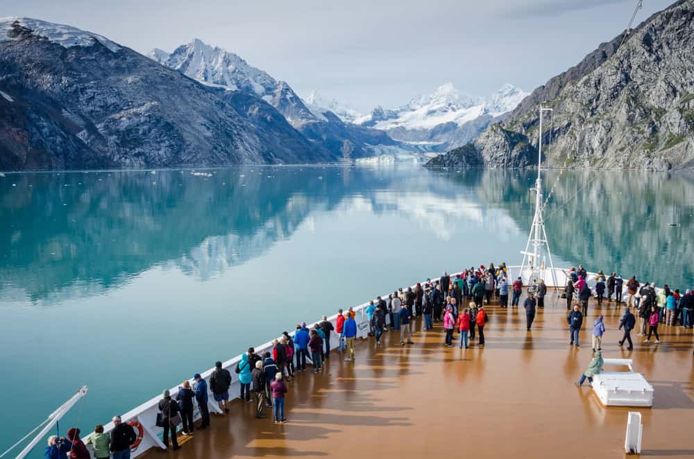 A large group of people stands on a ship deck, viewing a serene landscape of mountains and a glacier reflected in calm water.