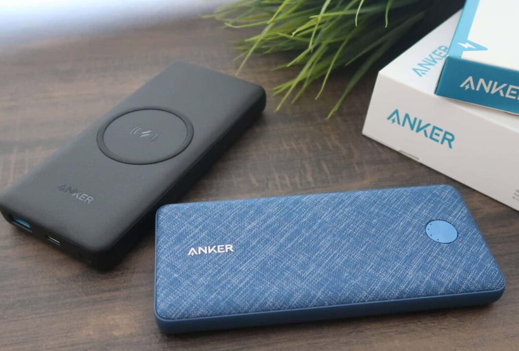 Two Anker power banks on a wooden surface; one is black with a wireless charging symbol, the other is blue with a fabric texture. An Anker box and green plant are in the background.