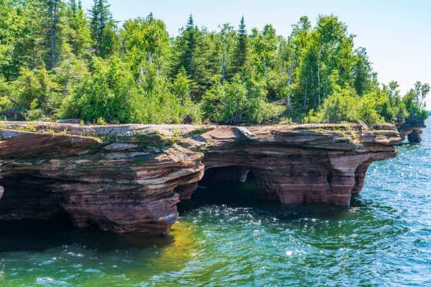 Rocky shoreline with trees, featuring sea caves and clear blue water under a sunny sky.