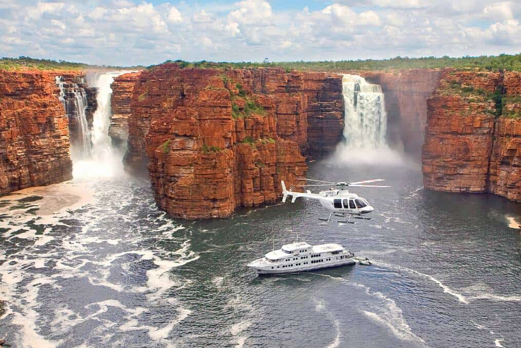 A helicopter flies over a yacht navigating the river near tall, rocky cliffs and waterfalls under a partly cloudy sky.