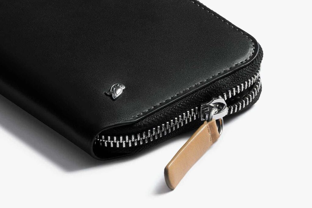 A black leather zippered wallet with a beige pull tab and a small silver logo on the corner.