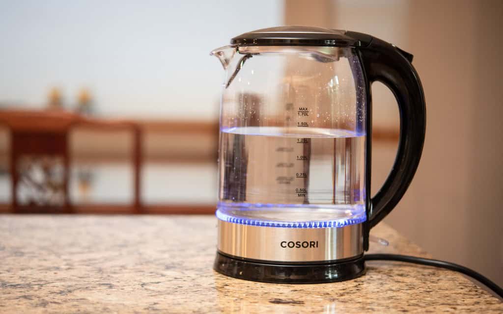 A glass electric kettle with a stainless steel base and black handle is filled with water, illuminated by a blue light, placed on a granite countertop.