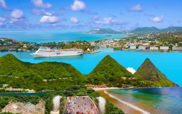 Aerial view of a coastal landscape with a cruise ship in a turquoise bay, surrounded by green hills and a town.