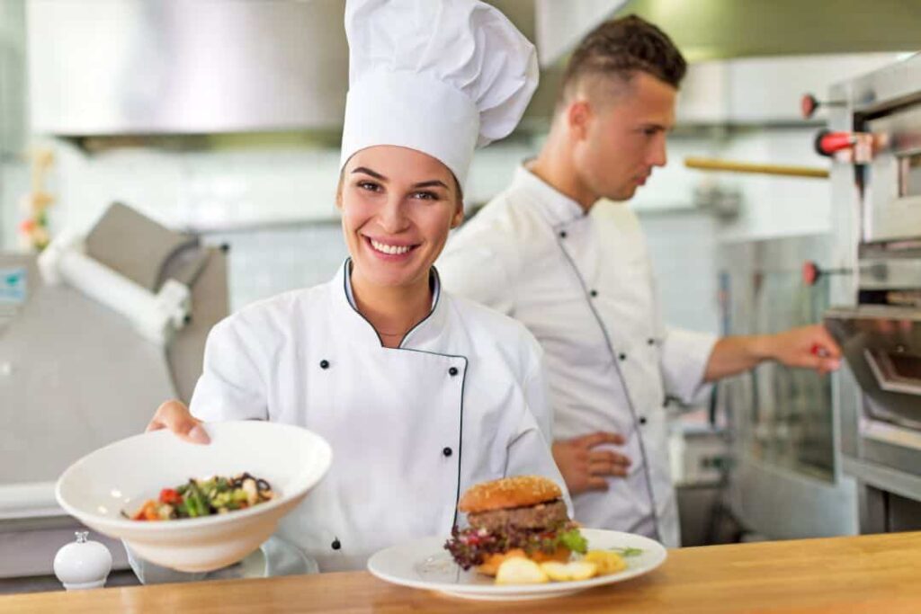 A smiling chef in a white uniform presents a dish of salad and a burger on a wooden counter in a restaurant kitchen. Another chef is working in the background.