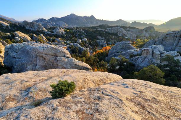 A rocky mountain landscape with boulders in the foreground, sparse vegetation, and distant peaks under a clear sky at sunset.