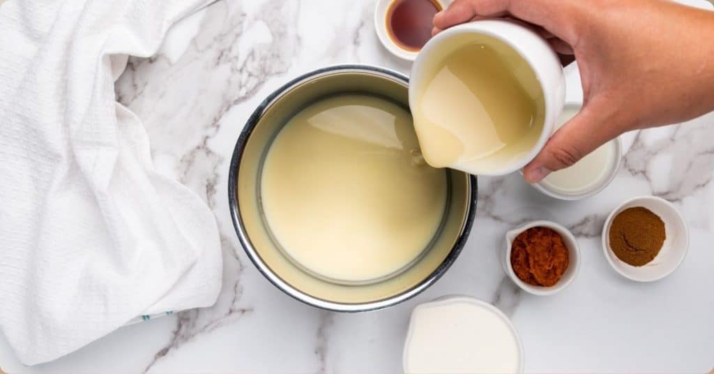 A person pours liquid from a white cup into a bowl on a marble countertop. Nearby are small bowls with brown powder, orange paste, and other ingredients, along with a white towel.