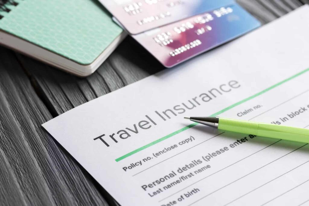 A travel insurance form with a green pen resting on it, two credit cards, and a notebook are placed on a textured wooden surface.