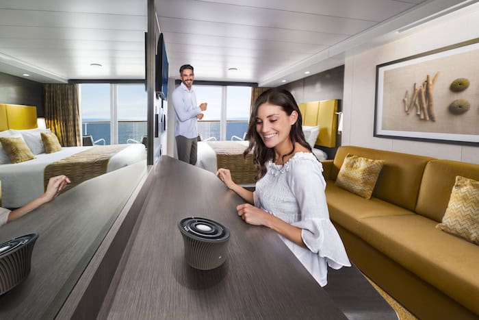 A woman sits at a desk with a small speaker, smiling. A man stands in the background near a bed, also smiling. The room has a modern design with a large mirror and ocean view.