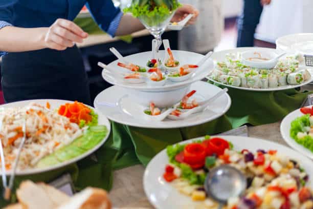 A buffet table featuring various dishes, including shrimp cocktails, sushi rolls, and fresh salads, with a person serving food in the background.