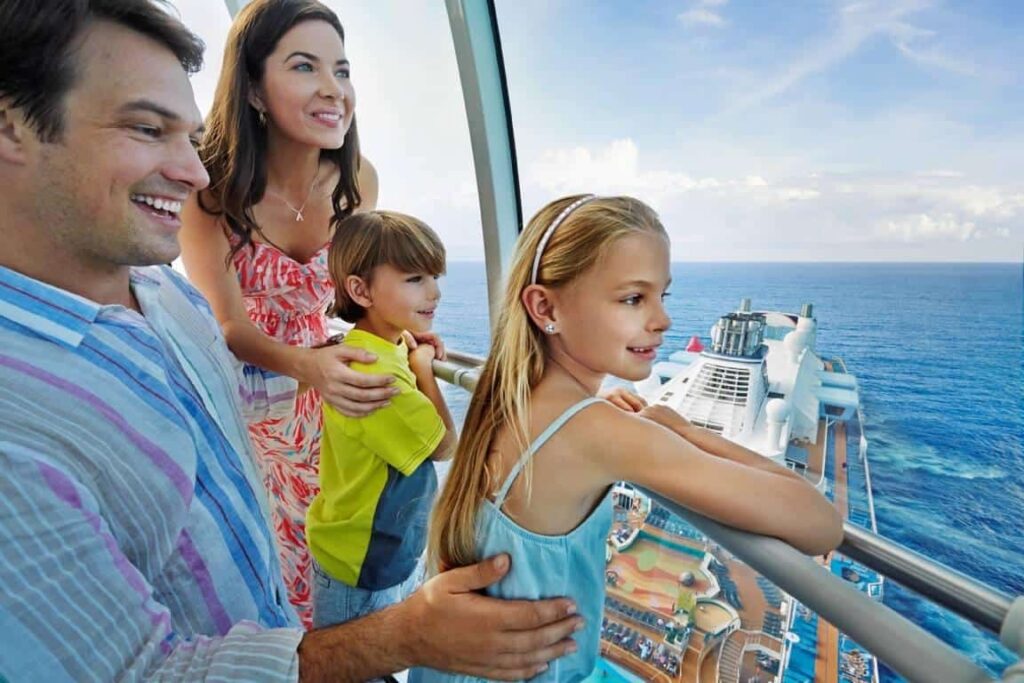 A family of four stands on a ship's deck, overlooking the ocean. They smile and look into the distance under a clear blue sky.