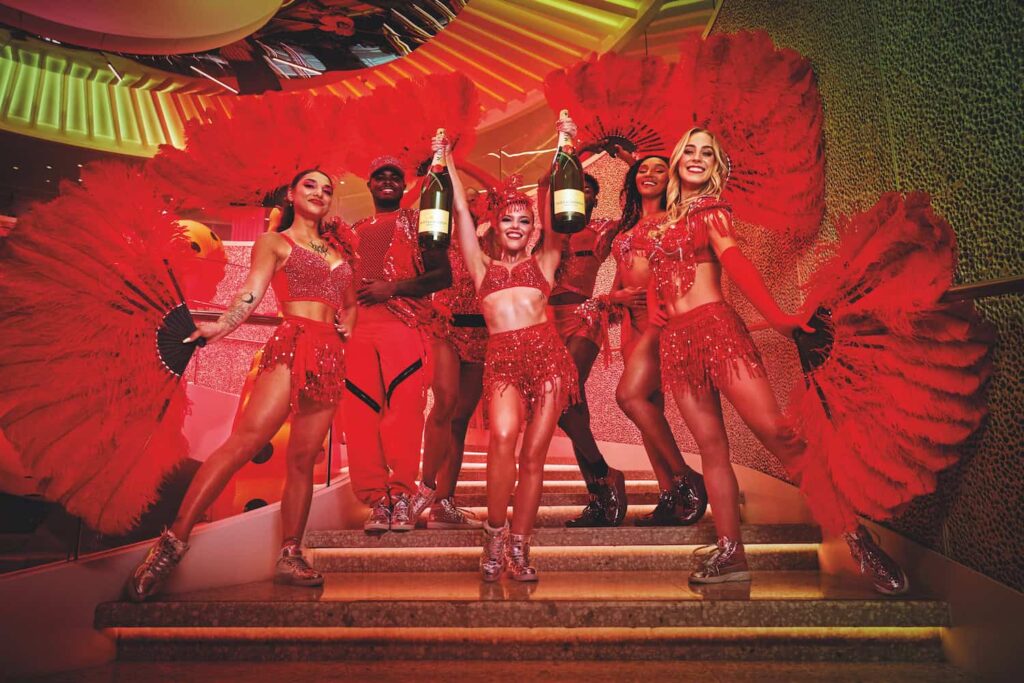 Performers in red costumes stand on stairs holding large feather fans and champagne bottles, smiling under vibrant lighting.