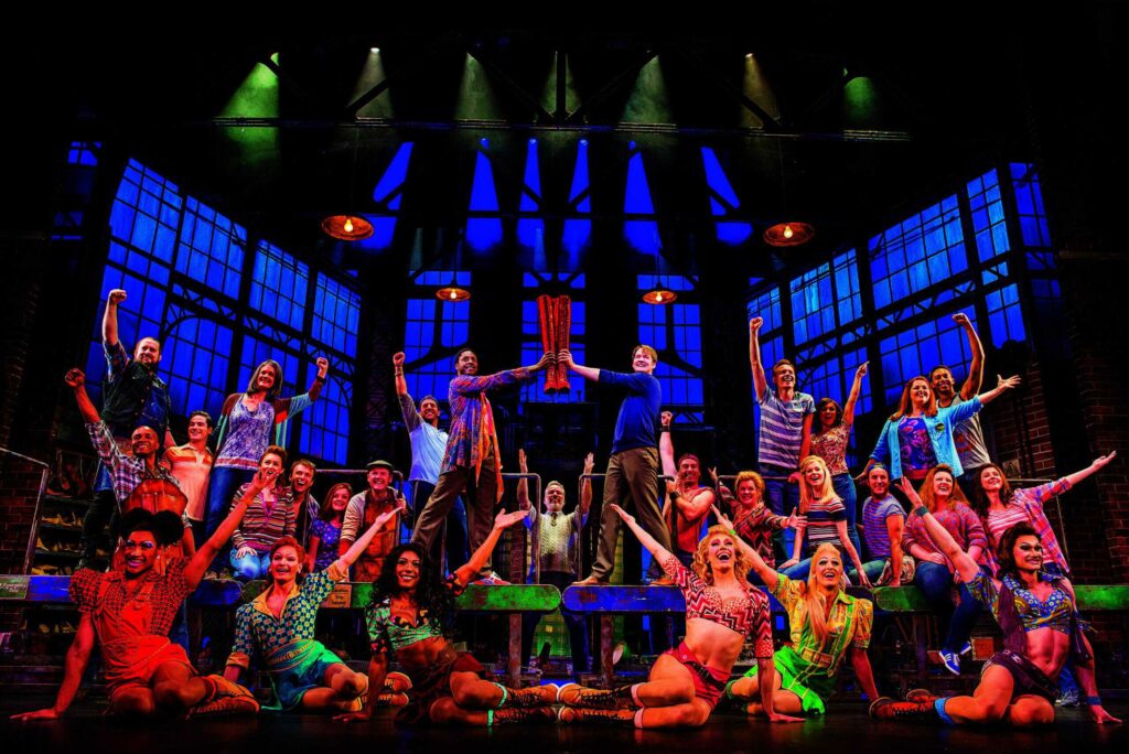 A vibrant theater cast poses energetically on stage, with two members prominently holding a large red boot above their heads. The background features industrial-themed set design.