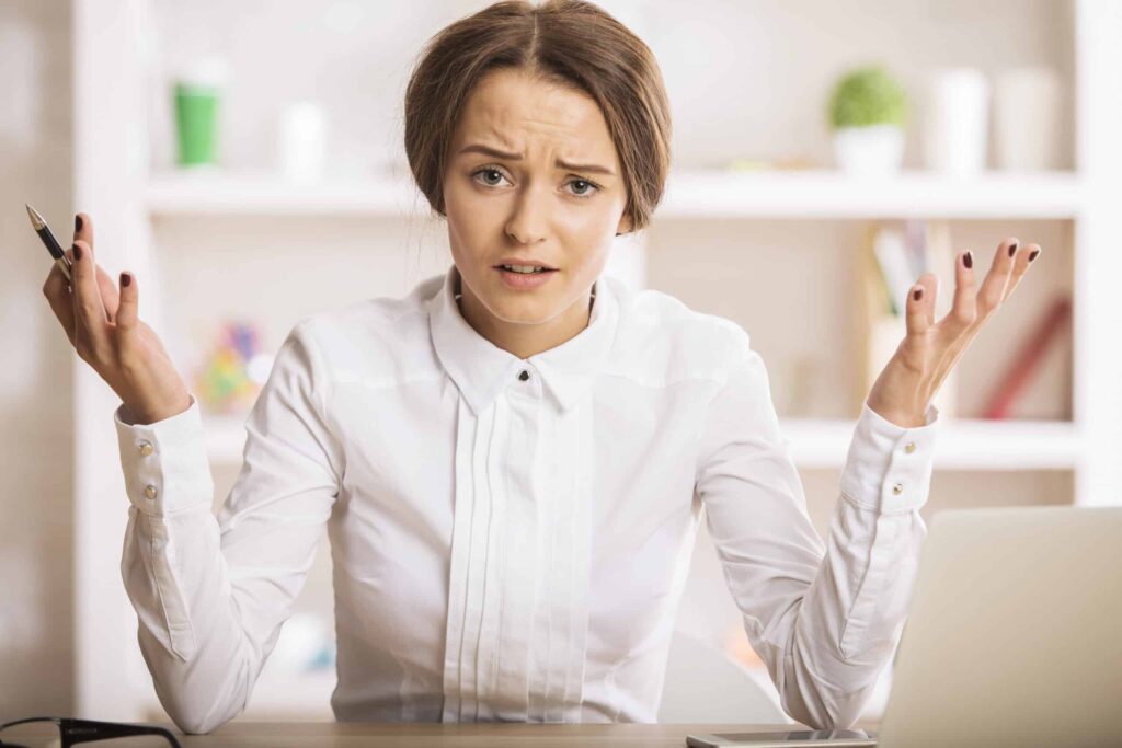 A person in a white shirt sits at a desk, holding a pen in one hand, looking puzzled with their arms raised. Shelves with decor are in the background.