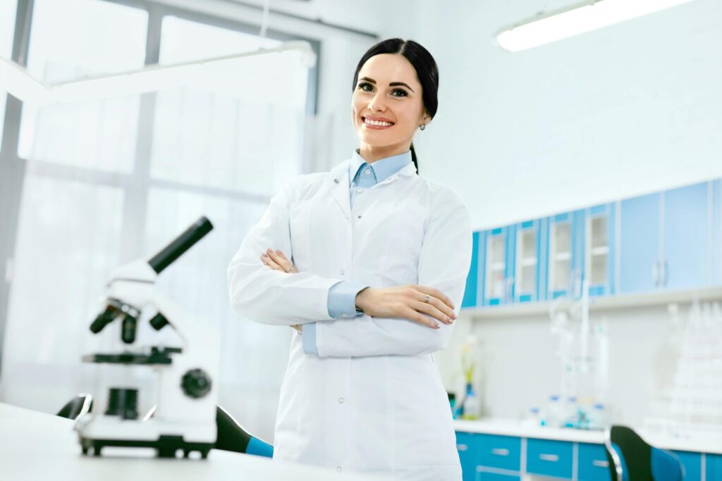 A scientist in a lab coat stands confidently with arms crossed in a laboratory, with a microscope on the desk nearby.