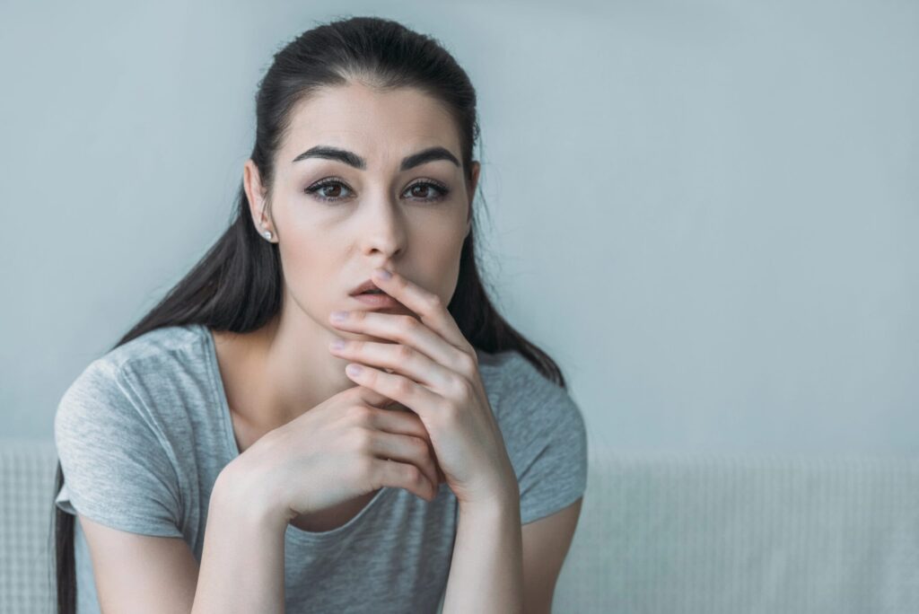 A person with long dark hair and a contemplative expression rests their chin on their fingers, wearing a gray shirt against a plain background.