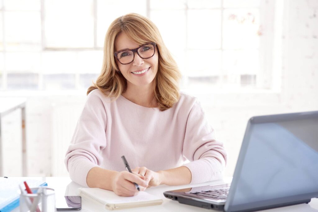 A woman with glasses sits at a desk, jotting down retirement strategies that work in her notebook, beside a laptop, in a brightly lit room.