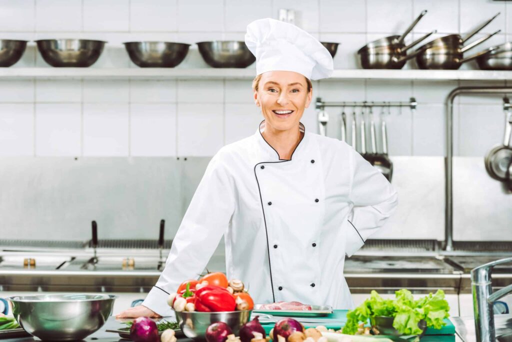 A chef in a white uniform and hat smiles while standing in a kitchen, with fresh vegetables and ingredients on the counter.