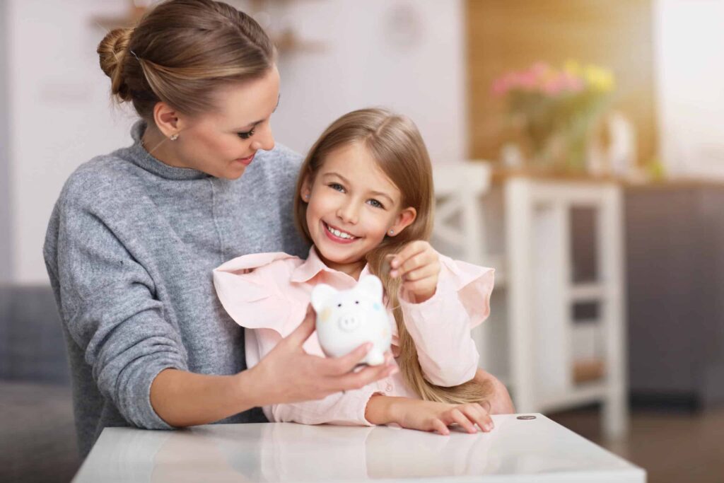 A woman and a child sit together at a table, smiling at a piggy bank, embracing generational financial habits with joy.