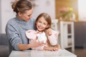 A woman and a child sit together at a table, smiling at a piggy bank, embracing generational financial habits with joy.