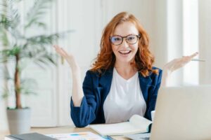A woman with red hair and glasses smiles and gestures with raised arms while sitting at a desk with a laptop and notebook. A plant is in the background.