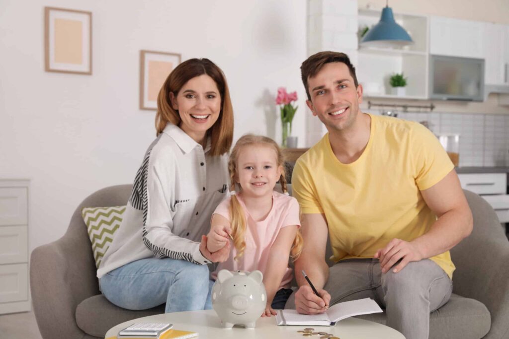 A family of three sits on a sofa in a living room. A girl is holding a coin above a piggy bank, while the parents smile at the camera. A notebook and coins are on the table.