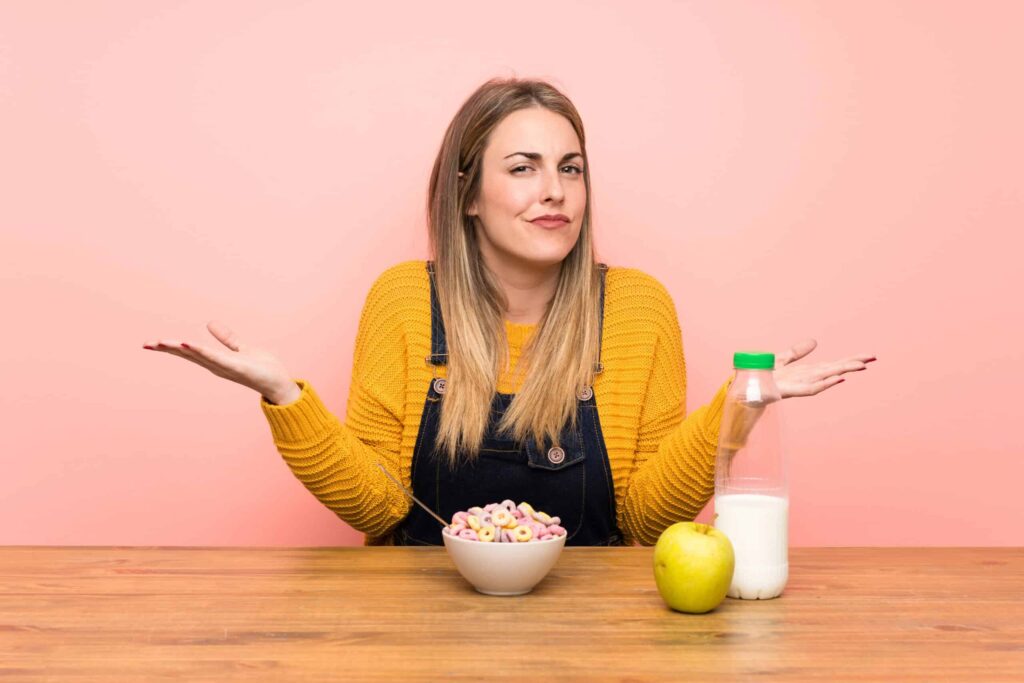 A woman in a yellow sweater shrugs at a table with a bowl of cereal, a milk bottle, and a green apple.