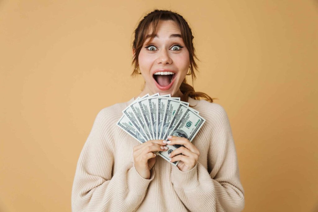 A person with a surprised expression holds several US dollar bills in front of a beige background, seemingly astonished by earnings from underrated side hustles that pay well.