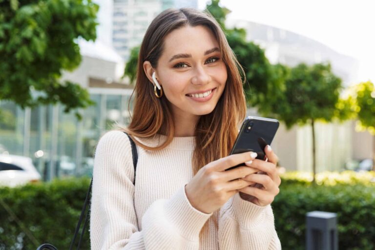 A woman in a white sweater stands outdoors, smiling and holding a smartphone with earbuds in her ears. Trees and buildings are visible in the background.