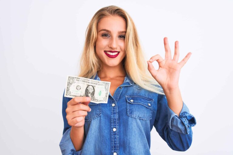Person in a denim shirt holding a one-dollar bill and making an "OK" gesture with their other hand.