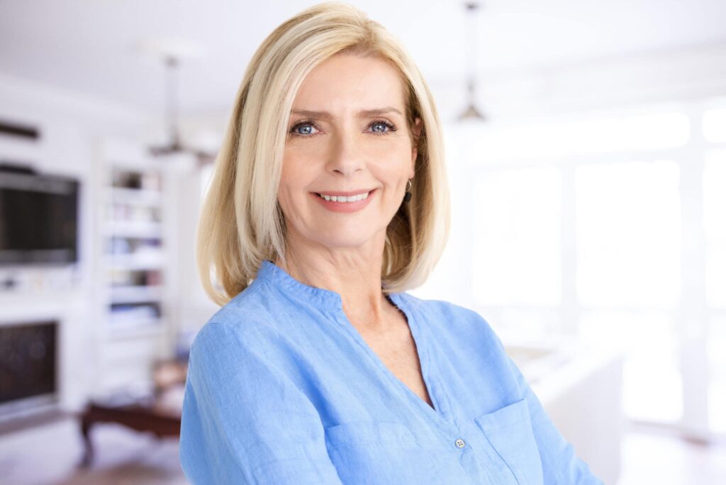A woman with blonde hair and a blue shirt smiles in a bright room.