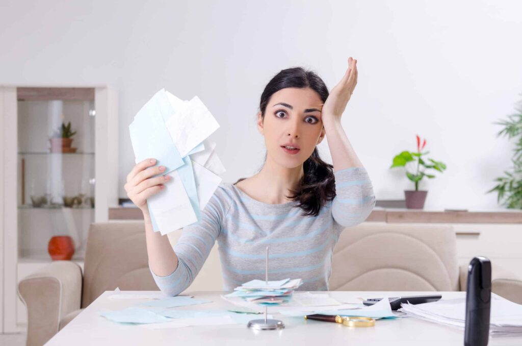 A woman sits at a table, holding several papers, looking surprised. The table has more papers, a pen, and a plant is visible in the background.