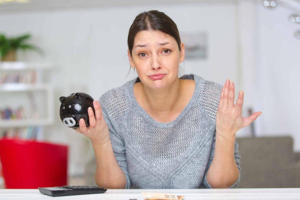 A woman in a gray sweater holds an empty black piggy bank in one hand and raises the other, looking concerned about why she's always broke. A calculator on the table suggests habits to break for better financial health.