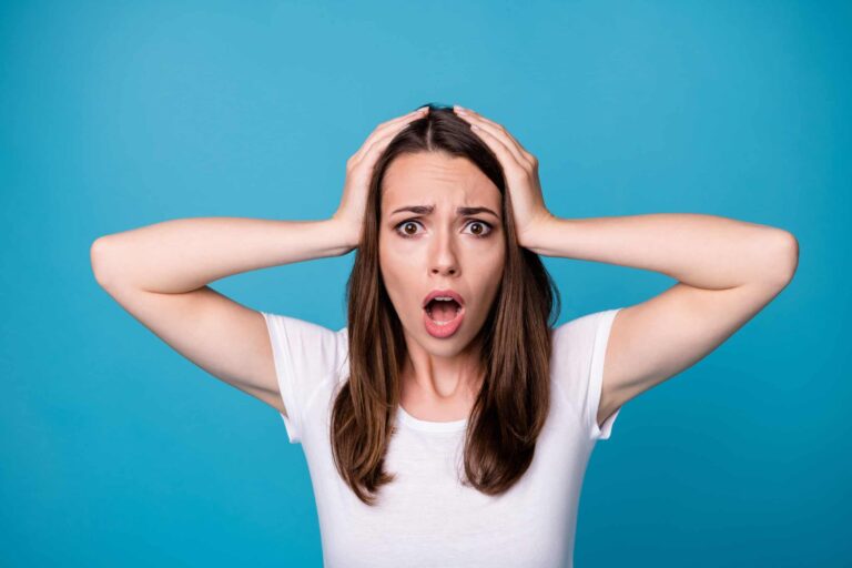 Woman with long brown hair in a white shirt stands against a blue background, holding her head with both hands and displaying a surprised expression.