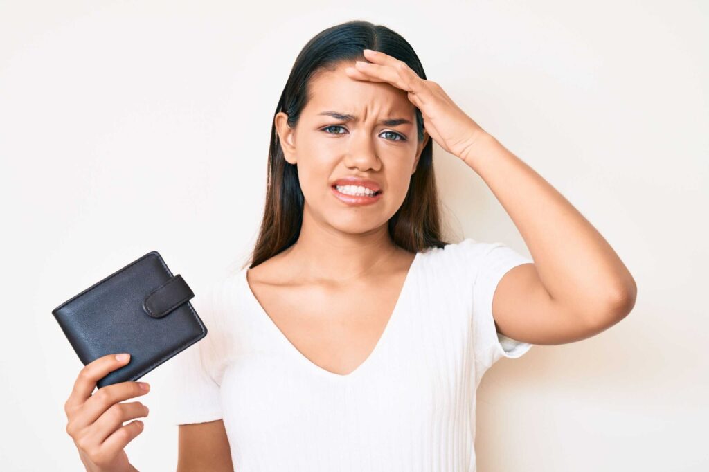 A woman in a white shirt holds a wallet and touches her forehead, appearing concerned or frustrated.