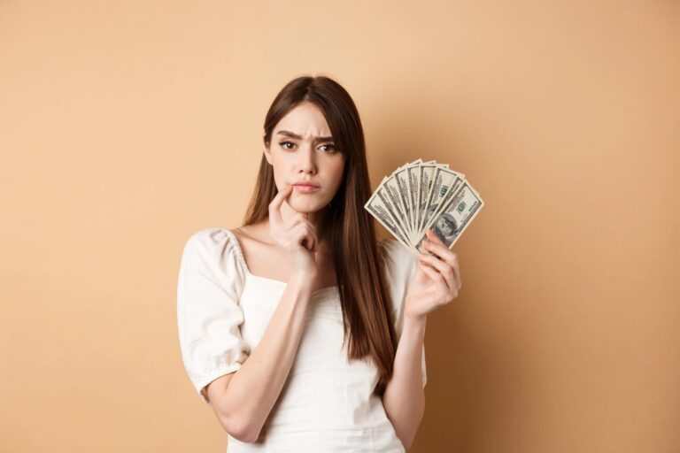 A woman in a white blouse holds a fan of dollar bills in one hand, looking thoughtful against a beige background.