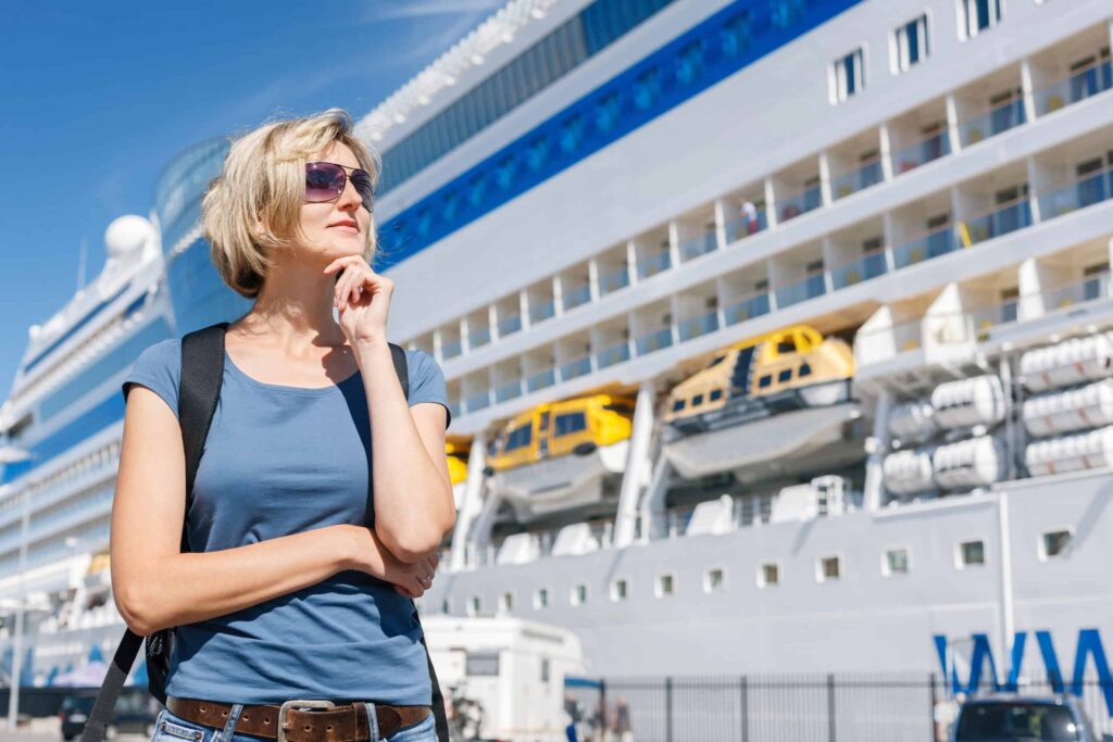 Woman in sunglasses stands in front of a large cruise ship, looking thoughtful.