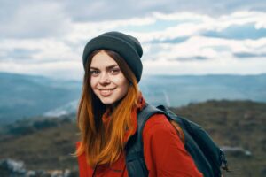 A person wearing a beanie and red jacket smiles while standing outdoors with a scenic mountain view in the background.