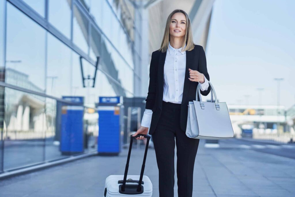 Woman in business attire pulling a suitcase and carrying a handbag outside an airport.