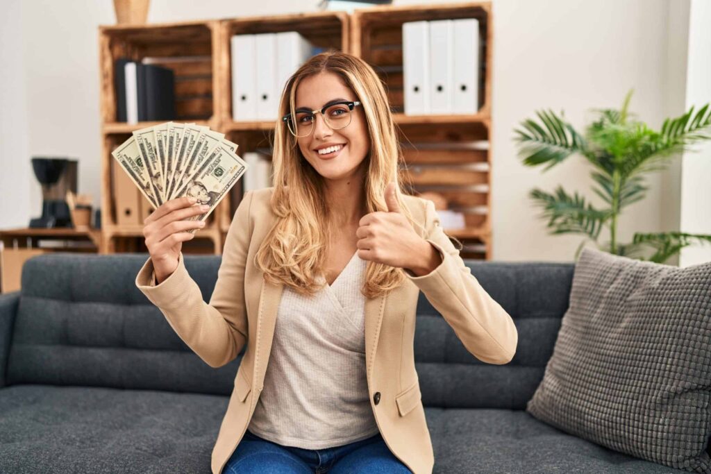 A woman sitting on a couch holds a fan of dollar bills in one hand and gives a thumbs-up with the other.