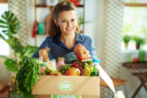A person smiling and holding a box filled with various groceries, including fruits, vegetables, and packaged goods, in a kitchen setting.