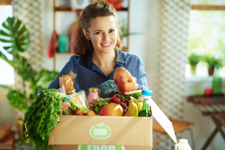 A person smiling and holding a box filled with various groceries, including fruits, vegetables, and packaged goods, in a kitchen setting.