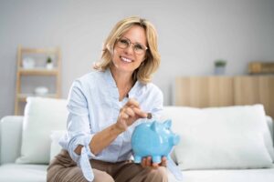 A woman with glasses smiles while placing a coin into a blue piggy bank, seated on a white couch in a living room.