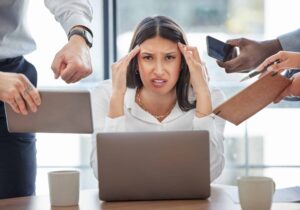 A stressed woman at a desk holds her head while surrounded by people's hands offering tablets, phones, and documents, emblematic of the ridiculous workplace rules overwhelming her daily life.
